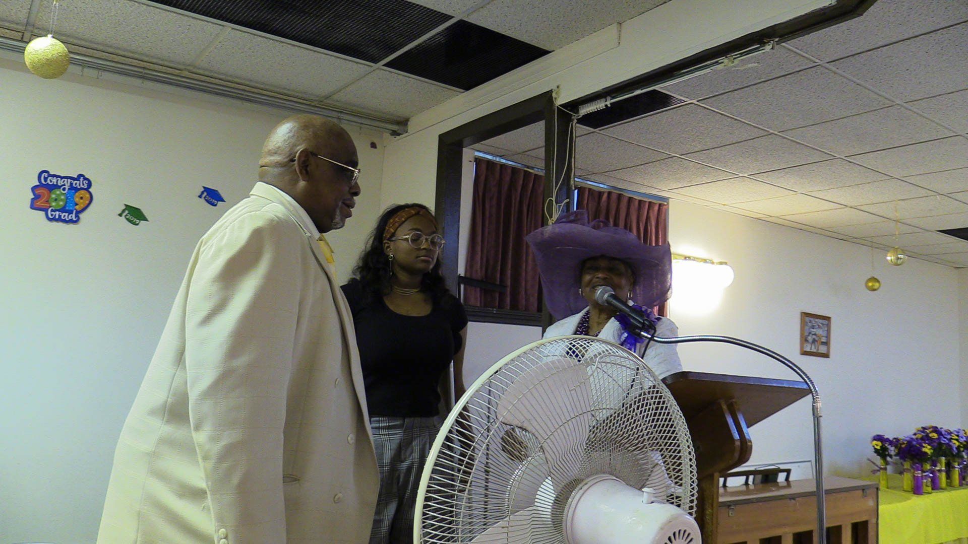 People standing near a podium in a small indoor room, with a fan in the foreground.