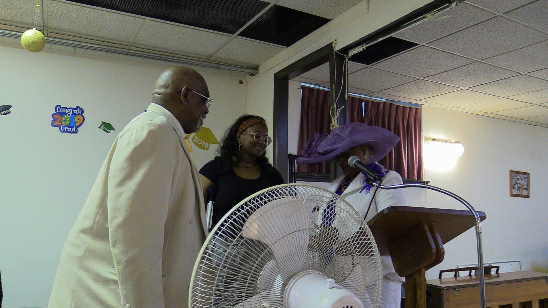 Two people stand near a lectern with a purple cloth in a small room with a large fan in the foreground.