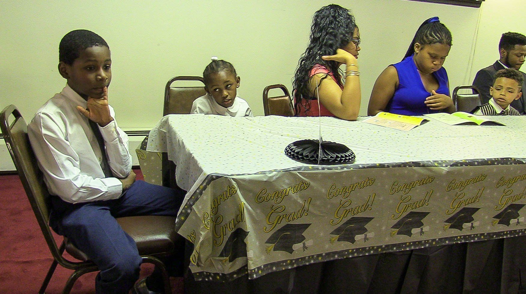 Several people sit around a dining table in a small room, with one person seated отдельно on the left.