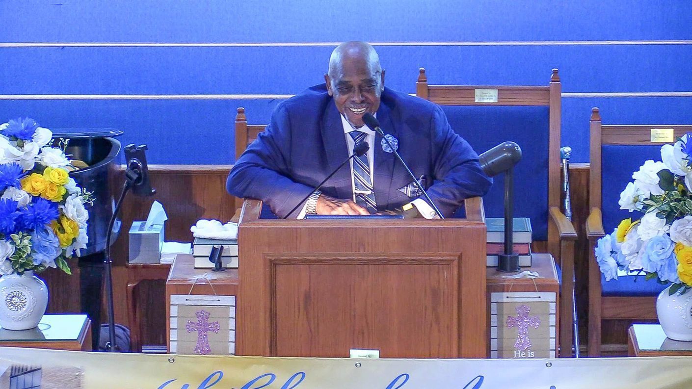 Pastor speaking at a wooden podium in a blue-walled church, flanked by floral arrangements.