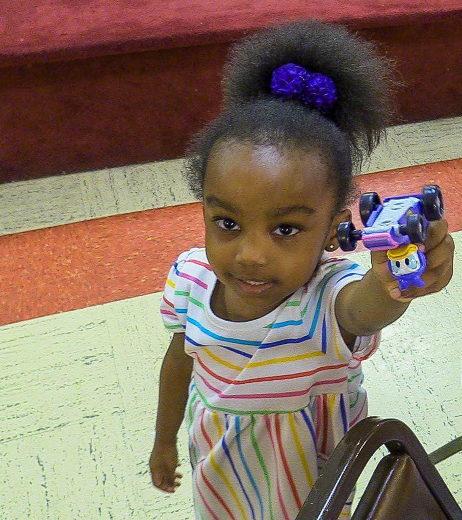 Child holding up a toy car indoors, looking up at the camera and smiling.