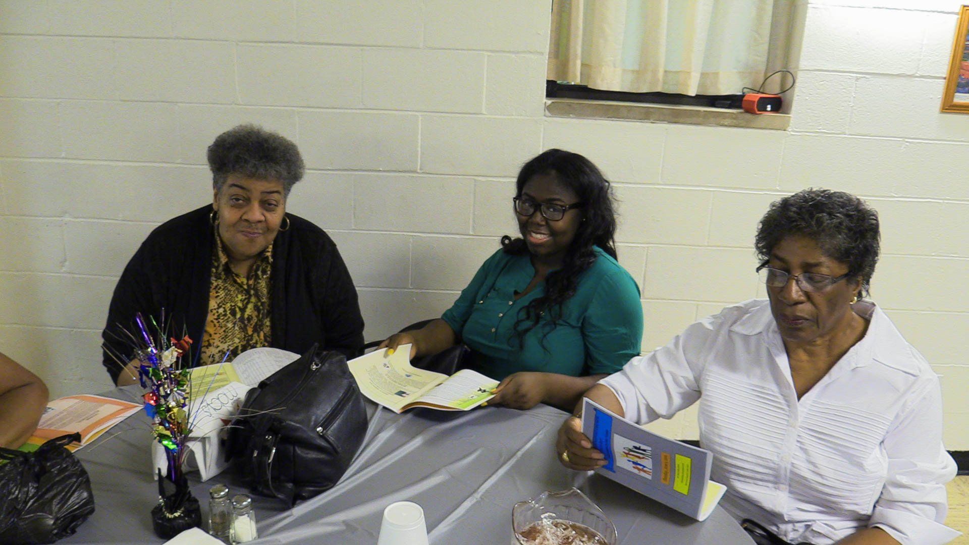 Three people seated at a table with papers and craft supplies in a classroom or meeting room.