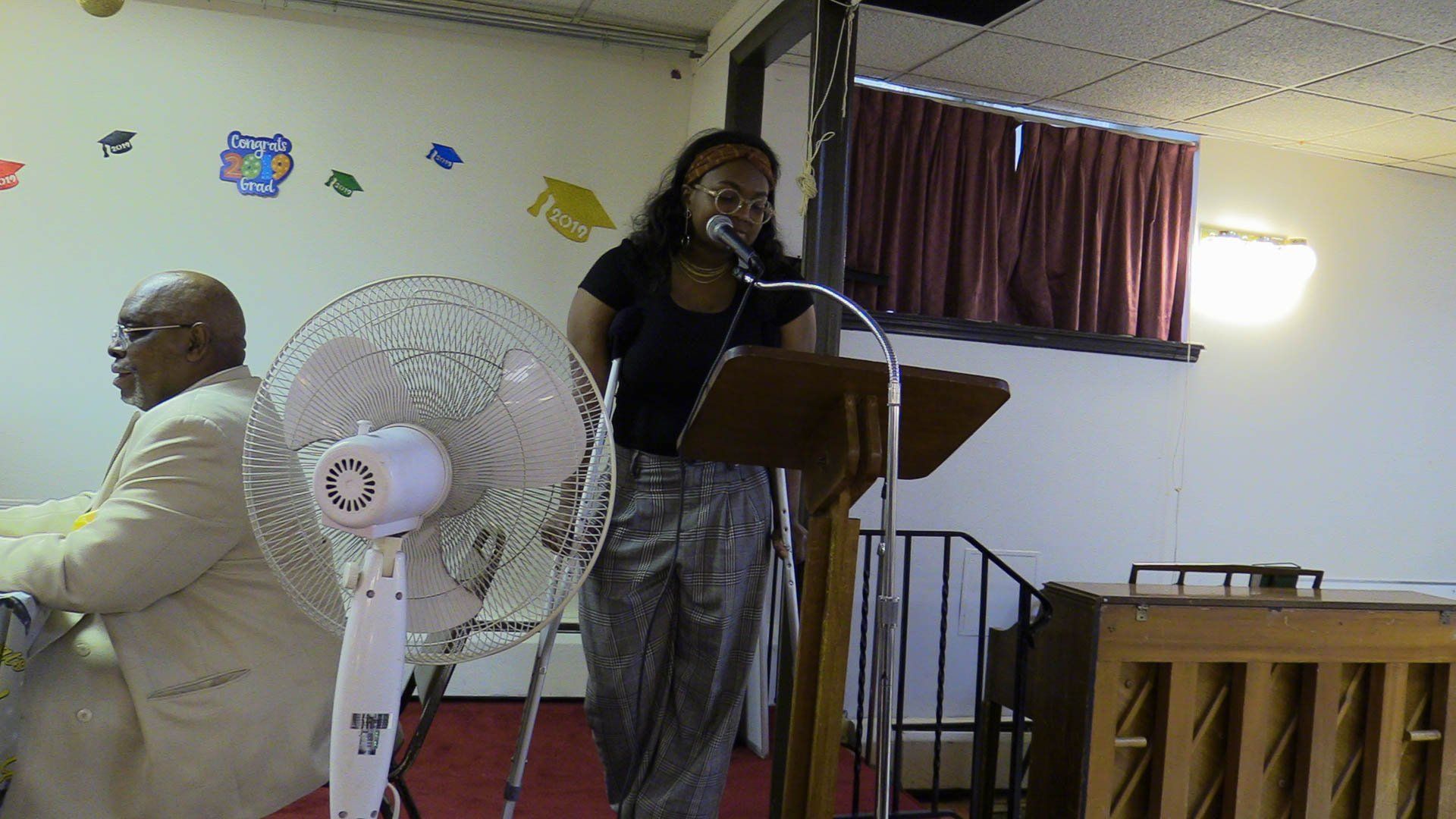 Woman speaking at a podium in a church, with a fan and piano nearby.