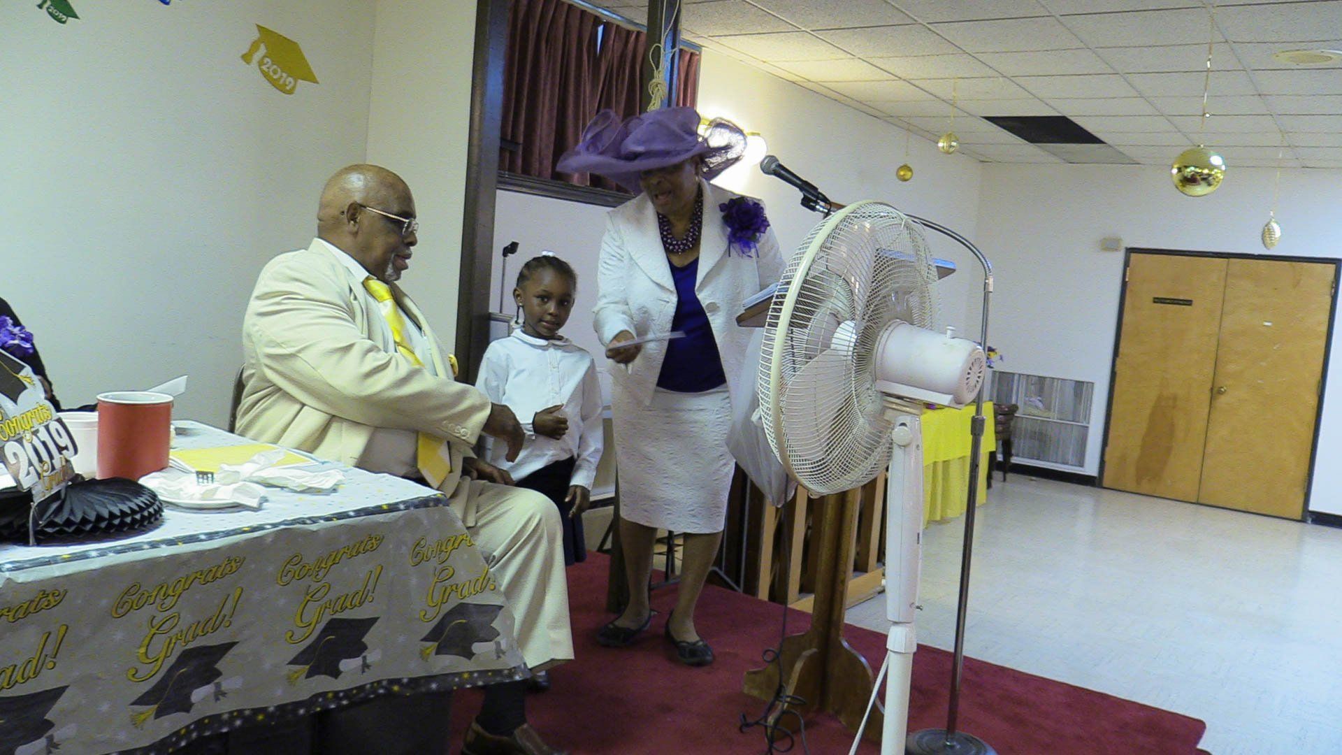 People gathered in a hall near a decorated table, with one person bending under a purple cloth.