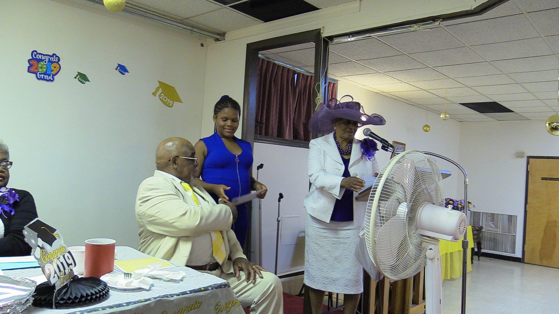 People gathered in a community room, with one person speaking beside a fan and table.