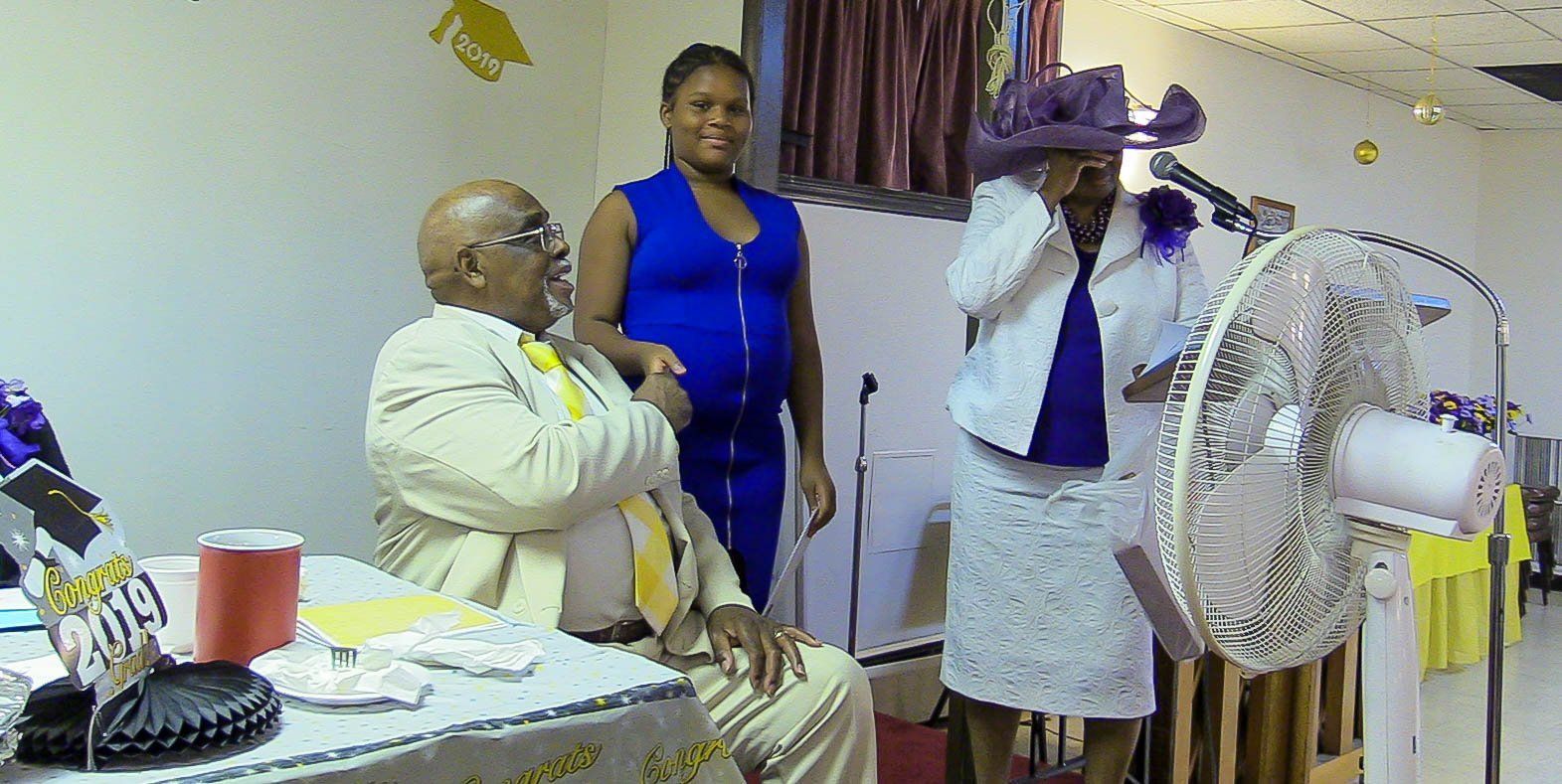 Three people standing and seated indoors at a table, one holding a microphone near a fan