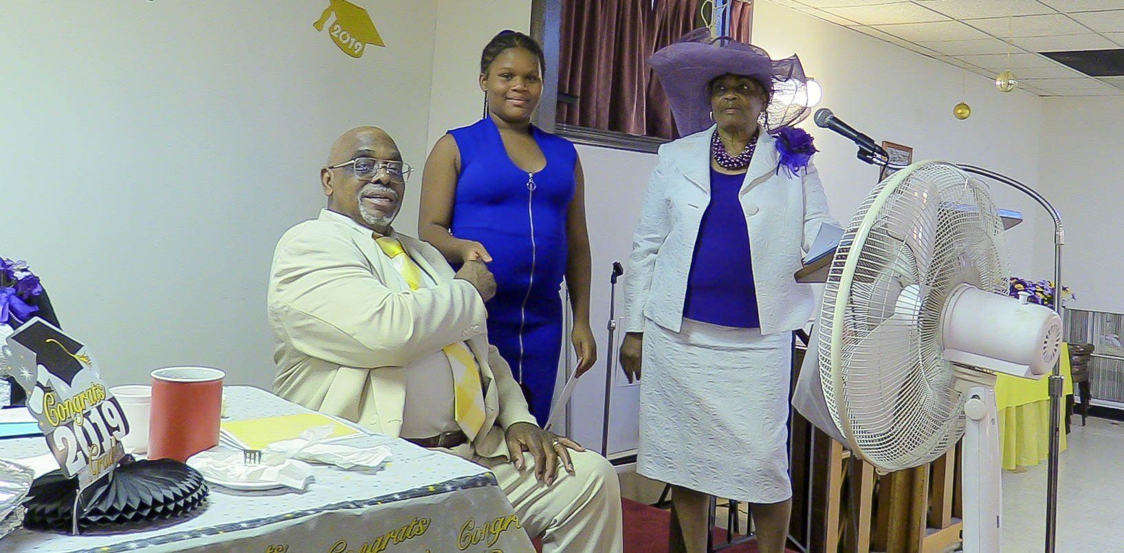 Three people posing indoors beside a table and standing fan, with one seated and two standing in blue and white outfits.