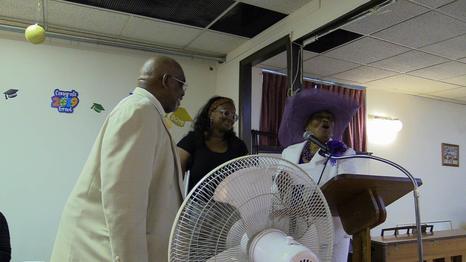People standing at a podium in a church, speaking into microphones beside a large fan.