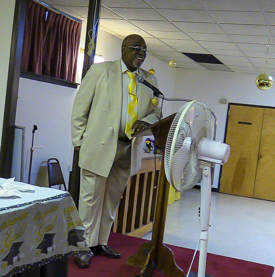 Man speaking at a podium in a hall, with a standing fan nearby and a red carpet on the floor