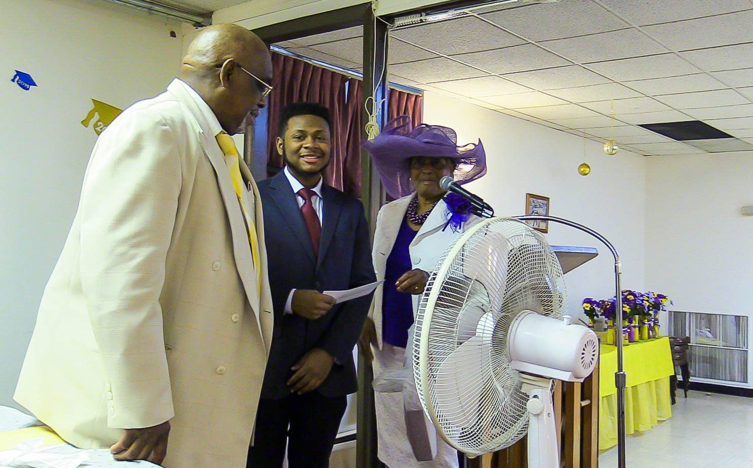Three people standing beside a fan on a stage in a decorated room, one speaking at a podium