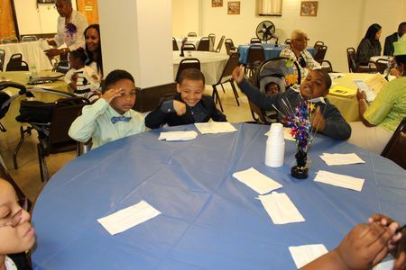 Children seated around a blue table at a classroom activity, with papers and a centerpiece on the table
