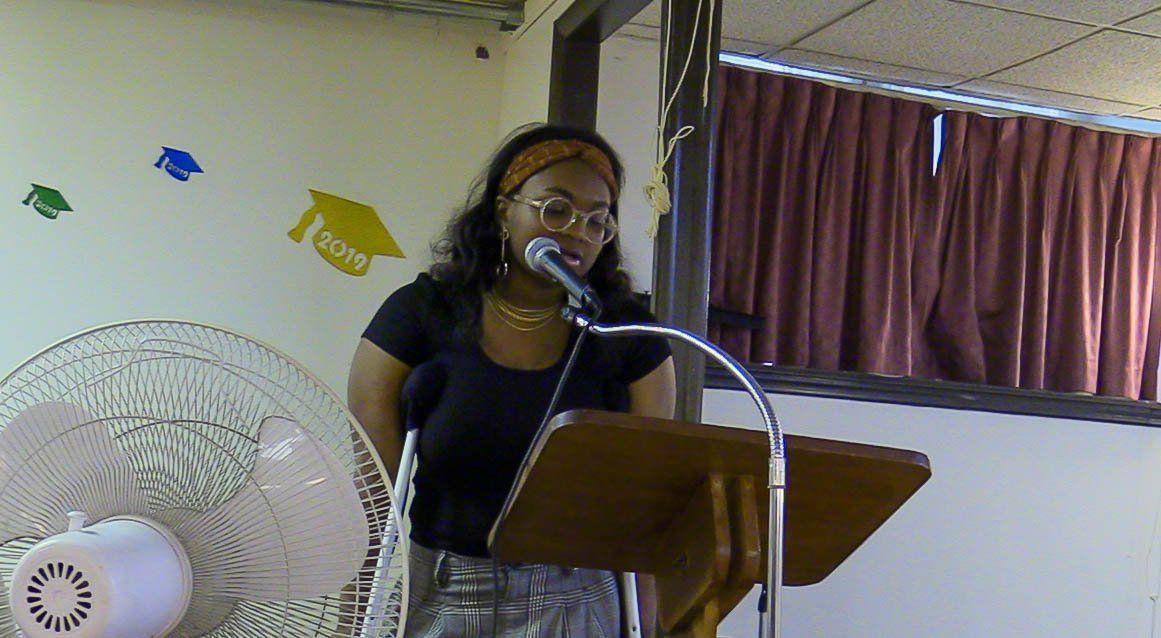 Woman speaking at a podium with a microphone in a small room, with curtains and wall decorations behind her