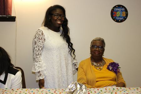 Two women at a decorated table, one standing in a white dress and one seated in a yellow outfit with a purple corsage.