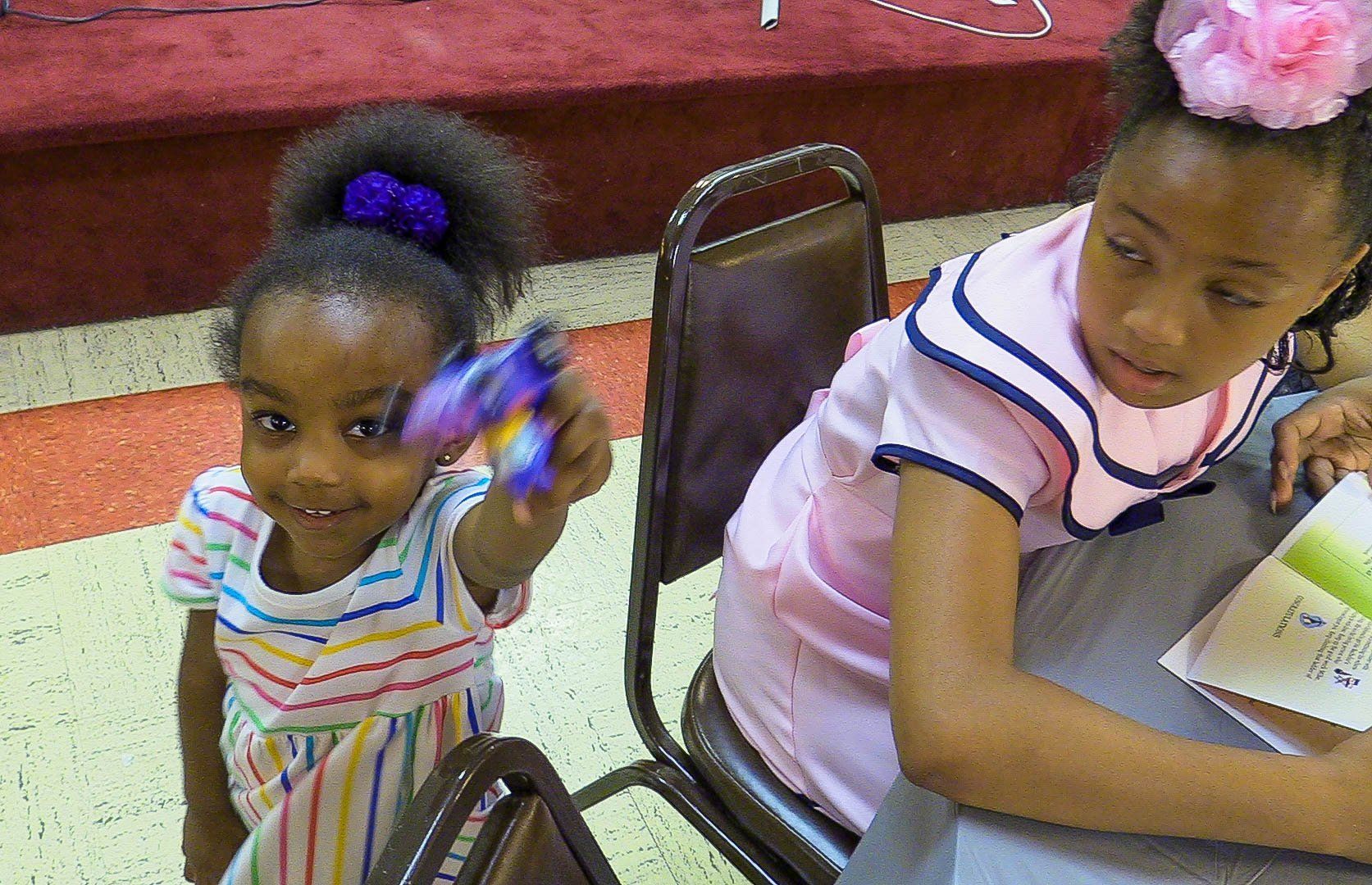 Two children seated indoors, one holding a colorful toy and the other reading a book.