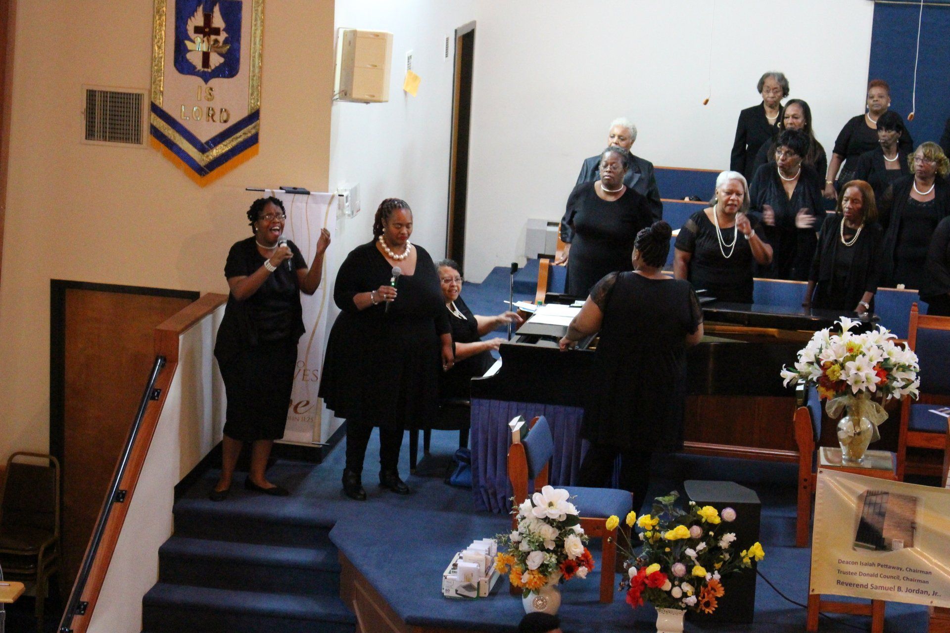 Choir singing in a church sanctuary, with singers standing near a piano and floral arrangements.