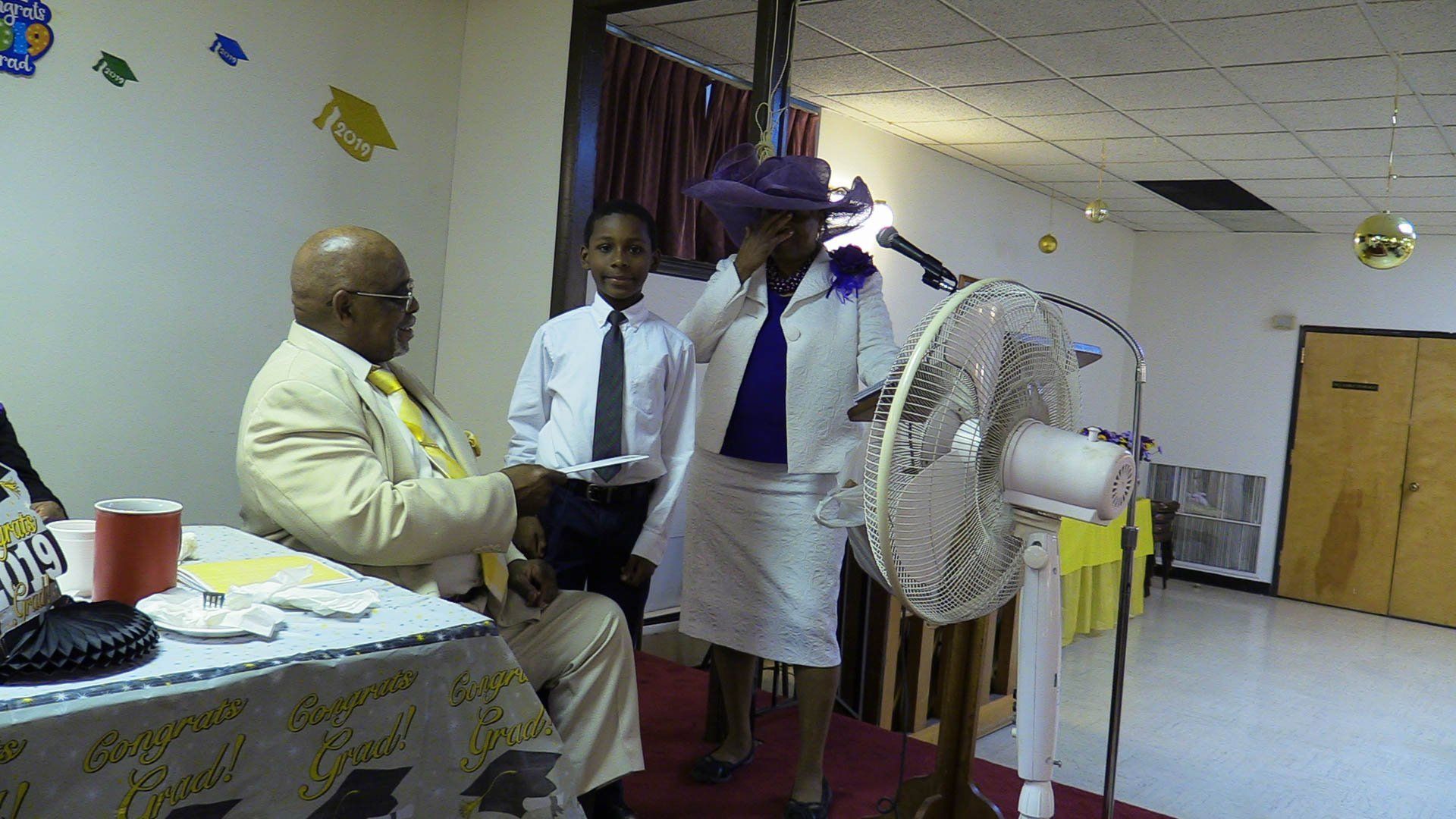 Man standing by a table facing a seated man at an indoor celebration with white decorations