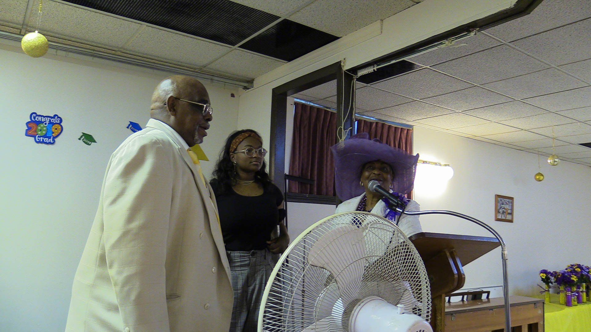 Three people standing at a podium in a church hall, with a fan in the foreground.