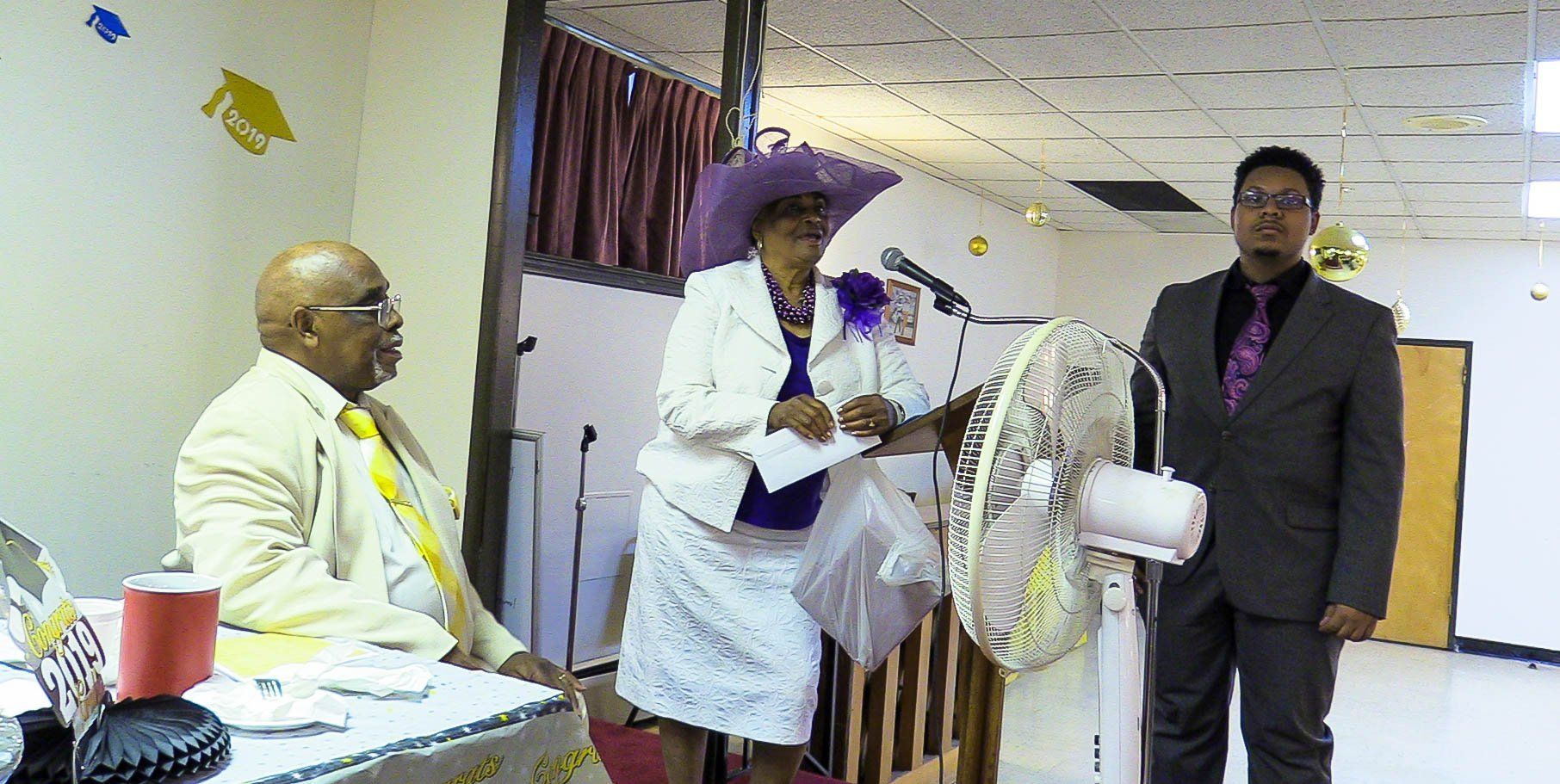 Three people at a party table near a large white fan in a decorated room