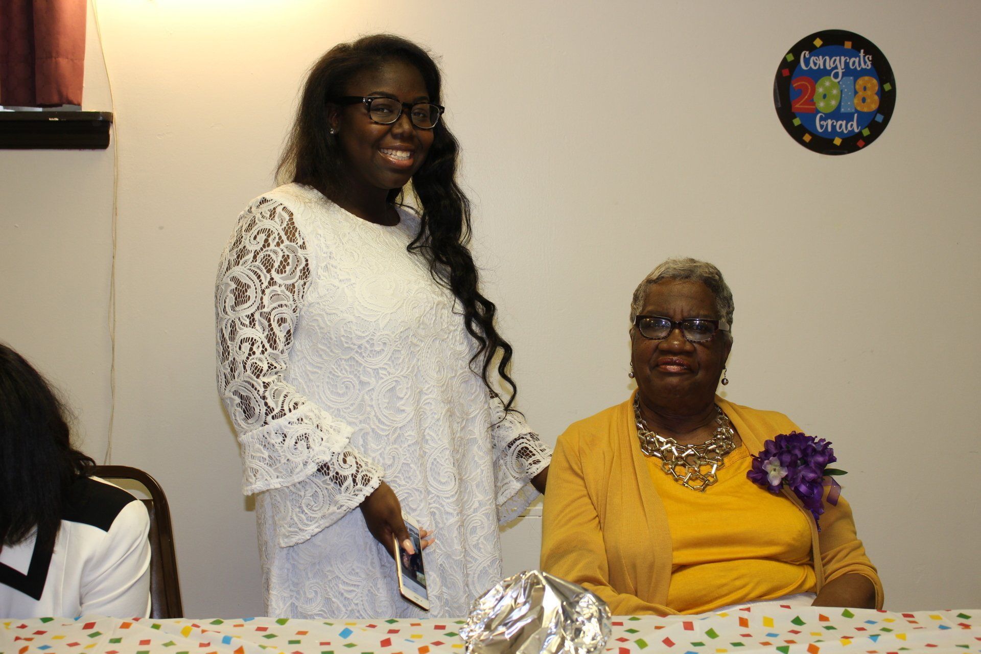 Two women at a decorated table, one standing and one seated, smiling indoors at a celebration