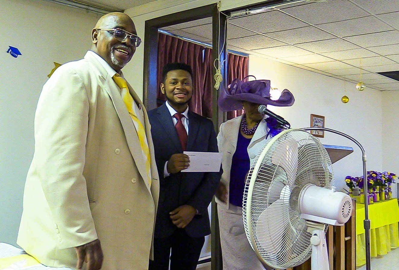 Three people at an indoor event, posing beside a large standing fan and holding a check.