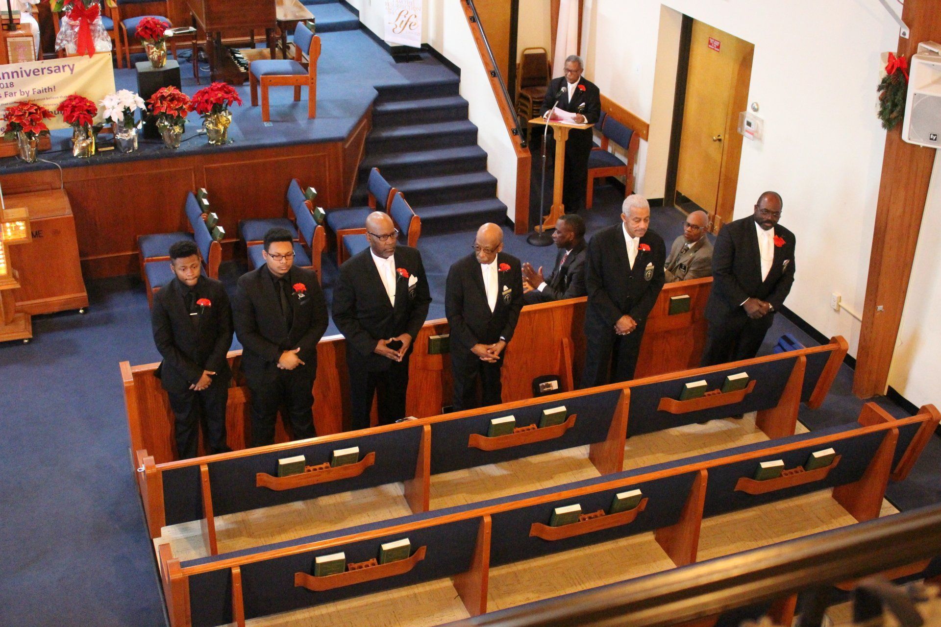 Men in dark suits standing in a church pew, with one man speaking at a lectern inside the sanctuary