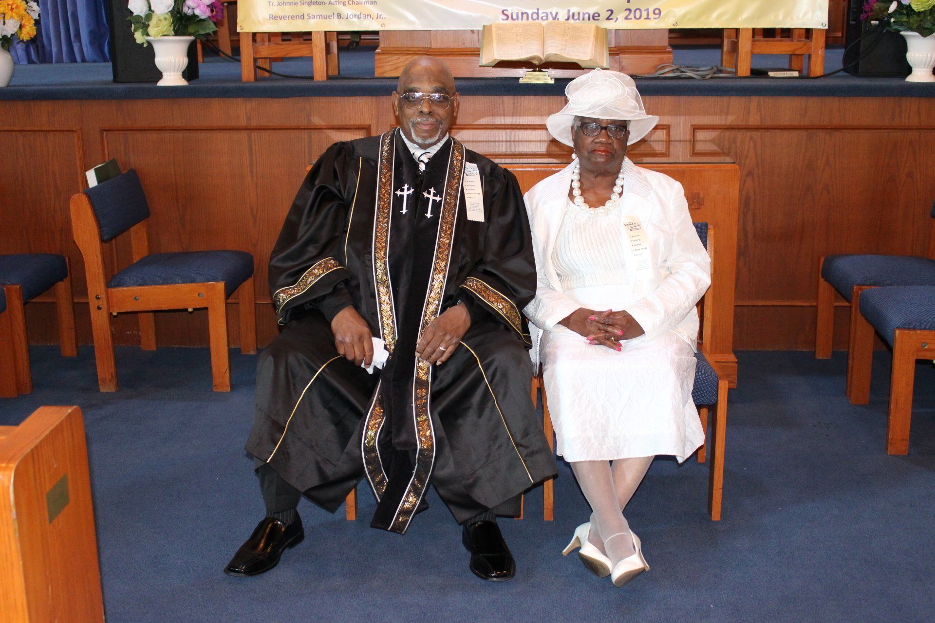 Two people seated on a church stage in formal attire, with blue carpet and wooden pews behind them