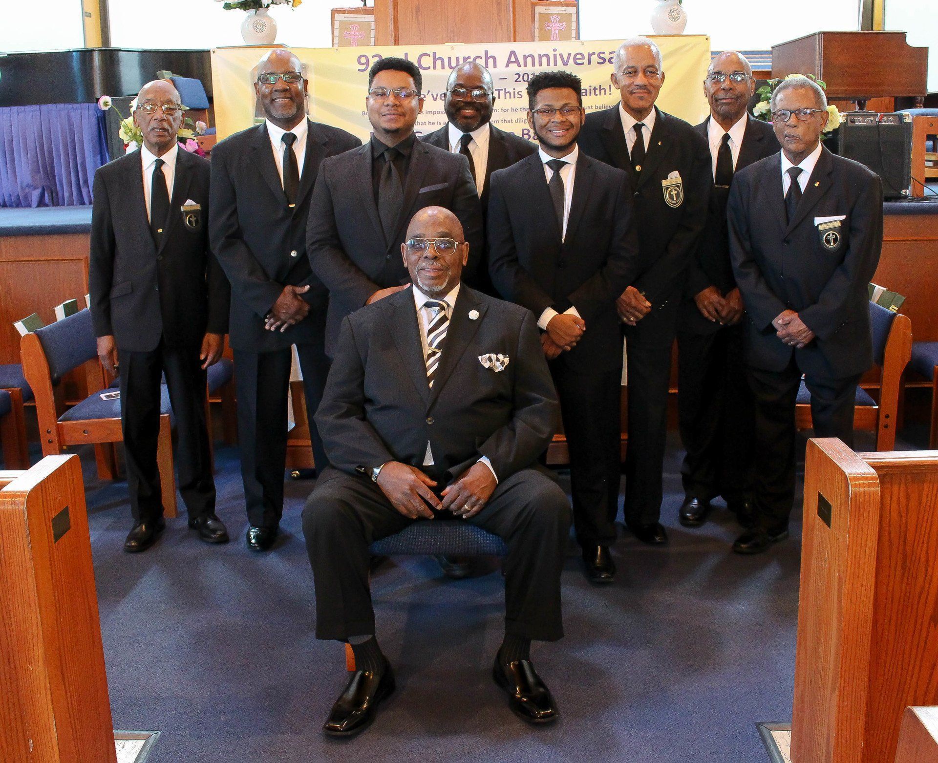 Group of men in dark suits posing inside a church, with one seated in front and pews behind them