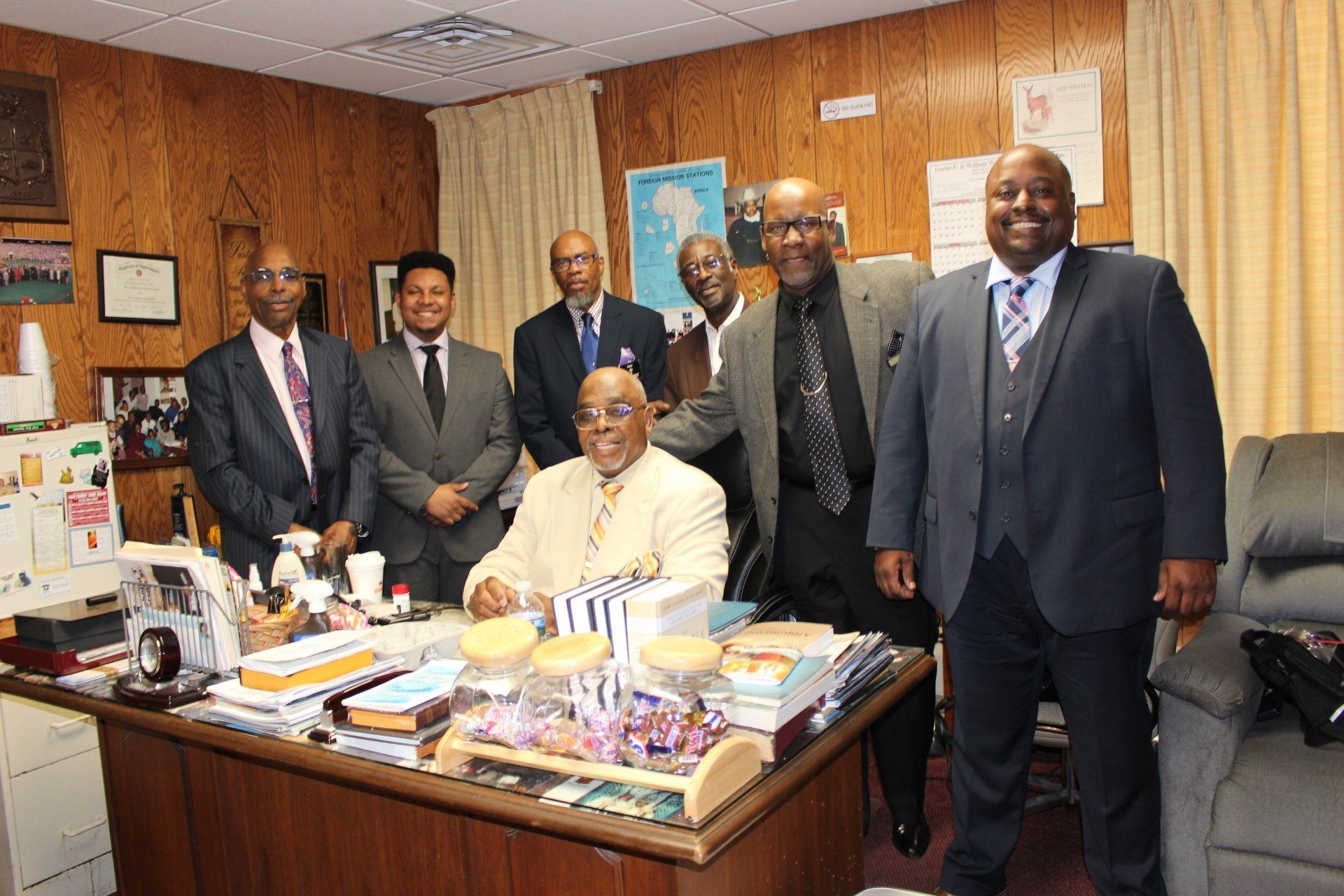 Group of people posing in an office, with a seated man behind a cluttered desk and wood-paneled walls.
