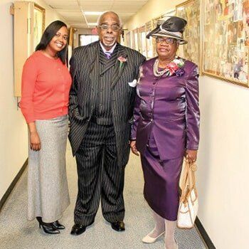Three people posing in a hallway, two in suits and one in a red top, all smiling at the camera.