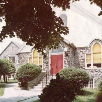 Stone church entrance with red doors, arched windows, and bushes under a tree canopy
