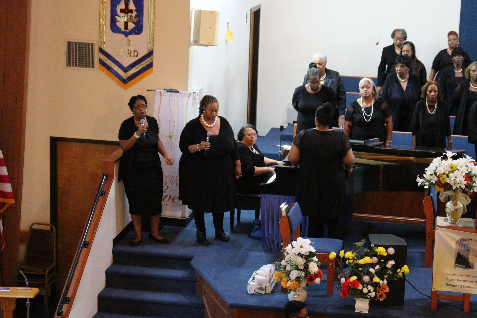 Choir performs in a church sanctuary beside a pulpit, with singers in black on a blue-carpeted stage.