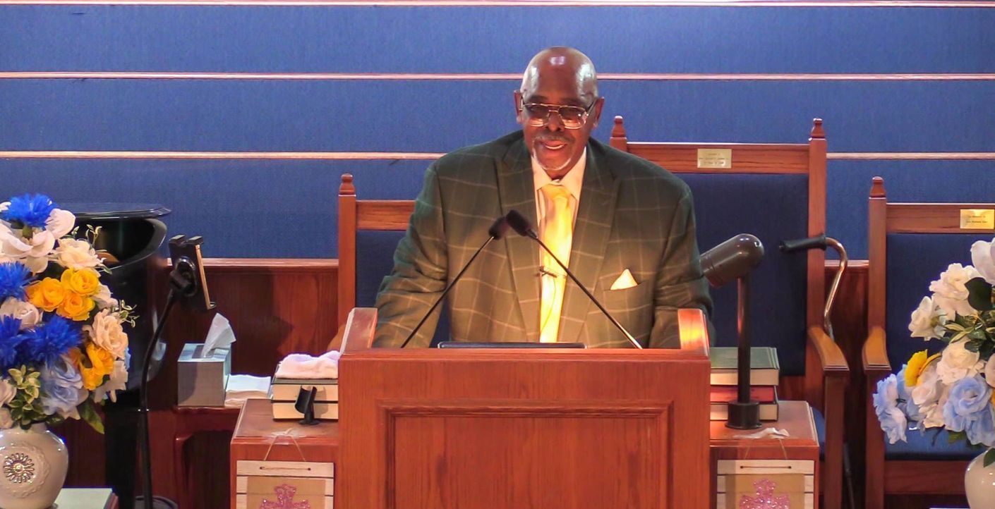 Man speaking at a wooden church podium with floral arrangements on both sides and blue walls behind him