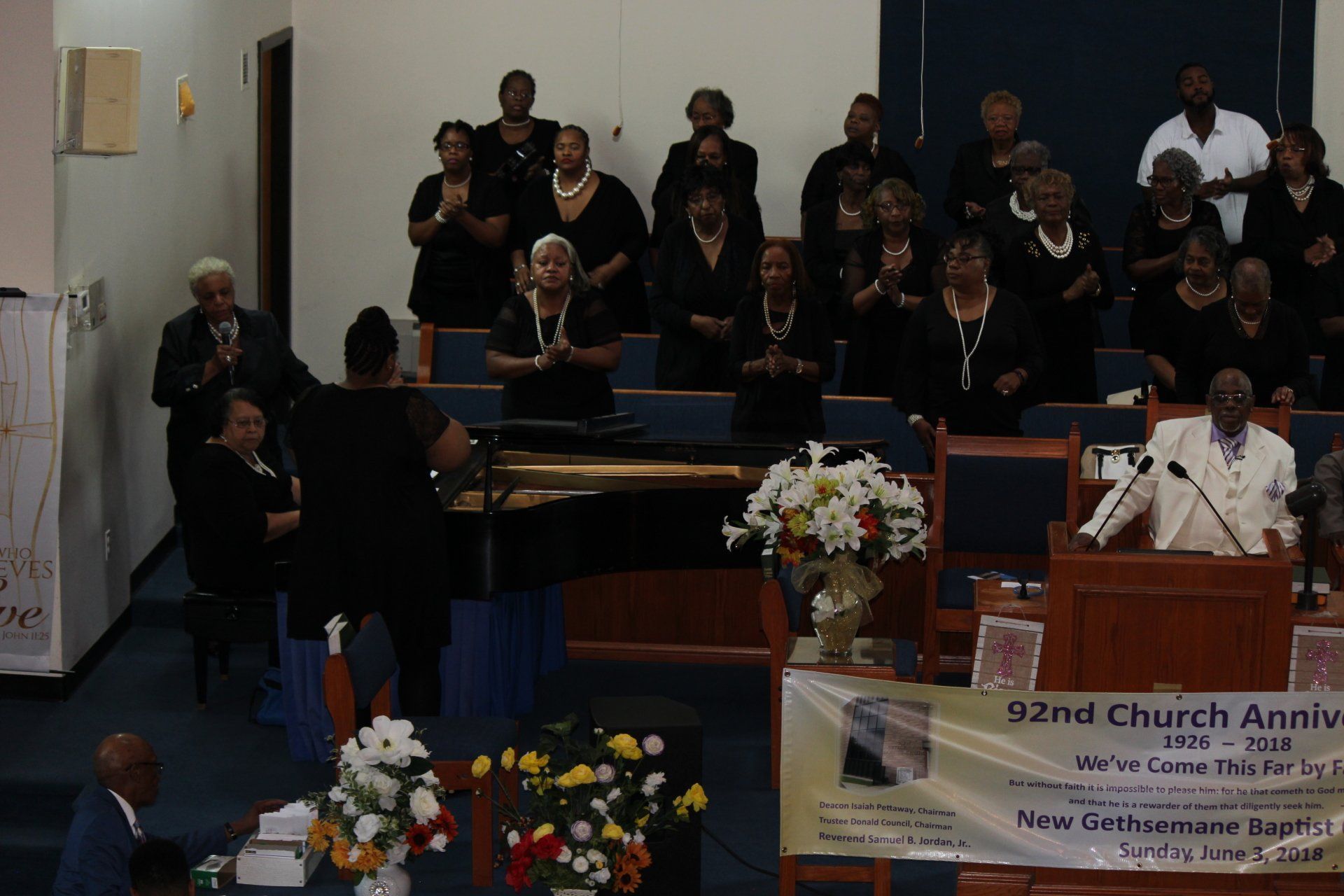 Choir in black robes singing in a church sanctuary beside a piano and floral arrangement