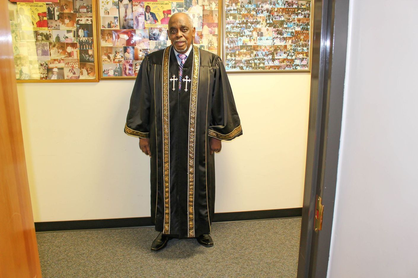 Pastor in a black robe with gold trim standing in a hallway before a bulletin board.