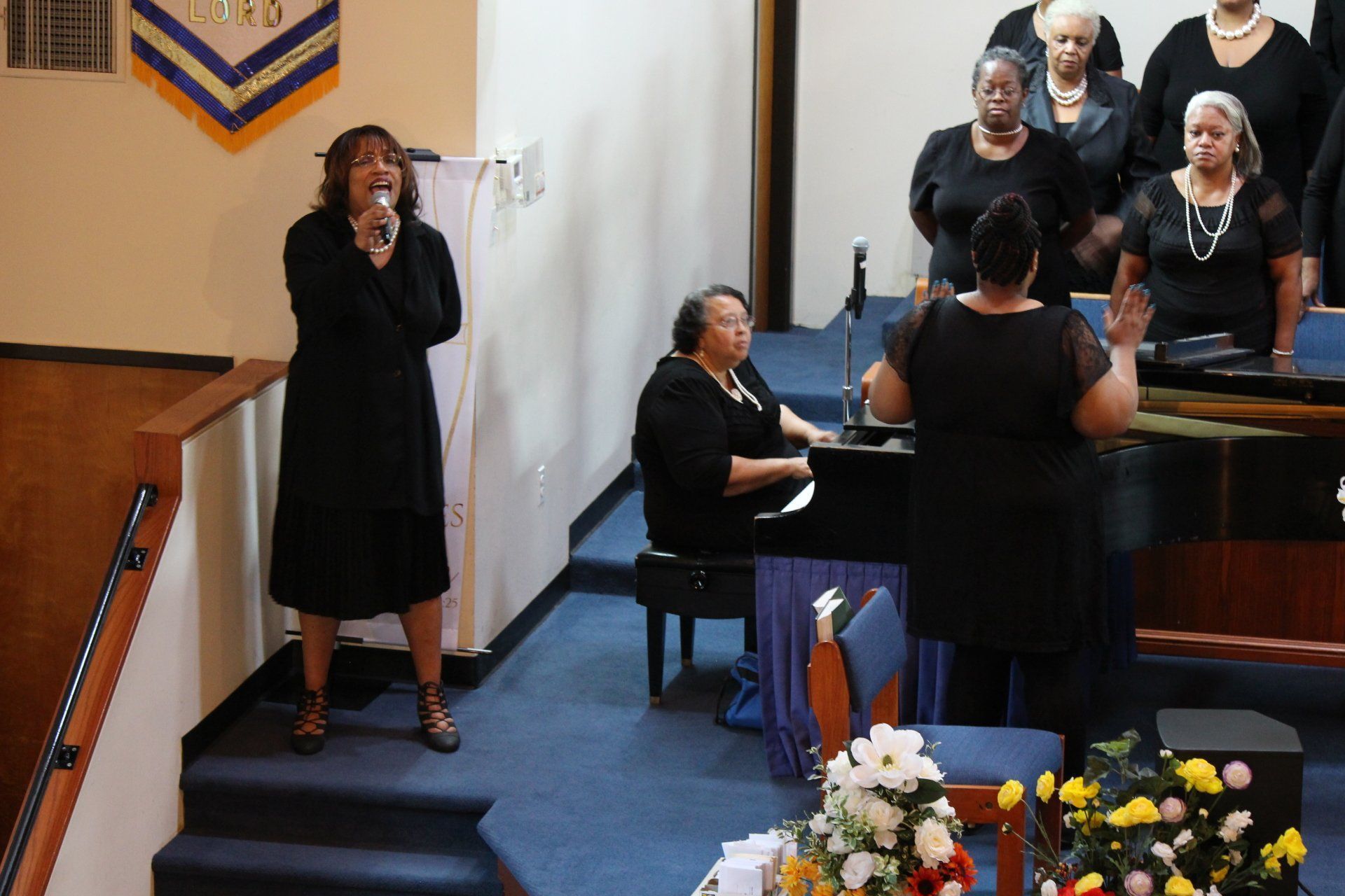 Woman singing at a funeral service beside a seated pianist and choir in black attire