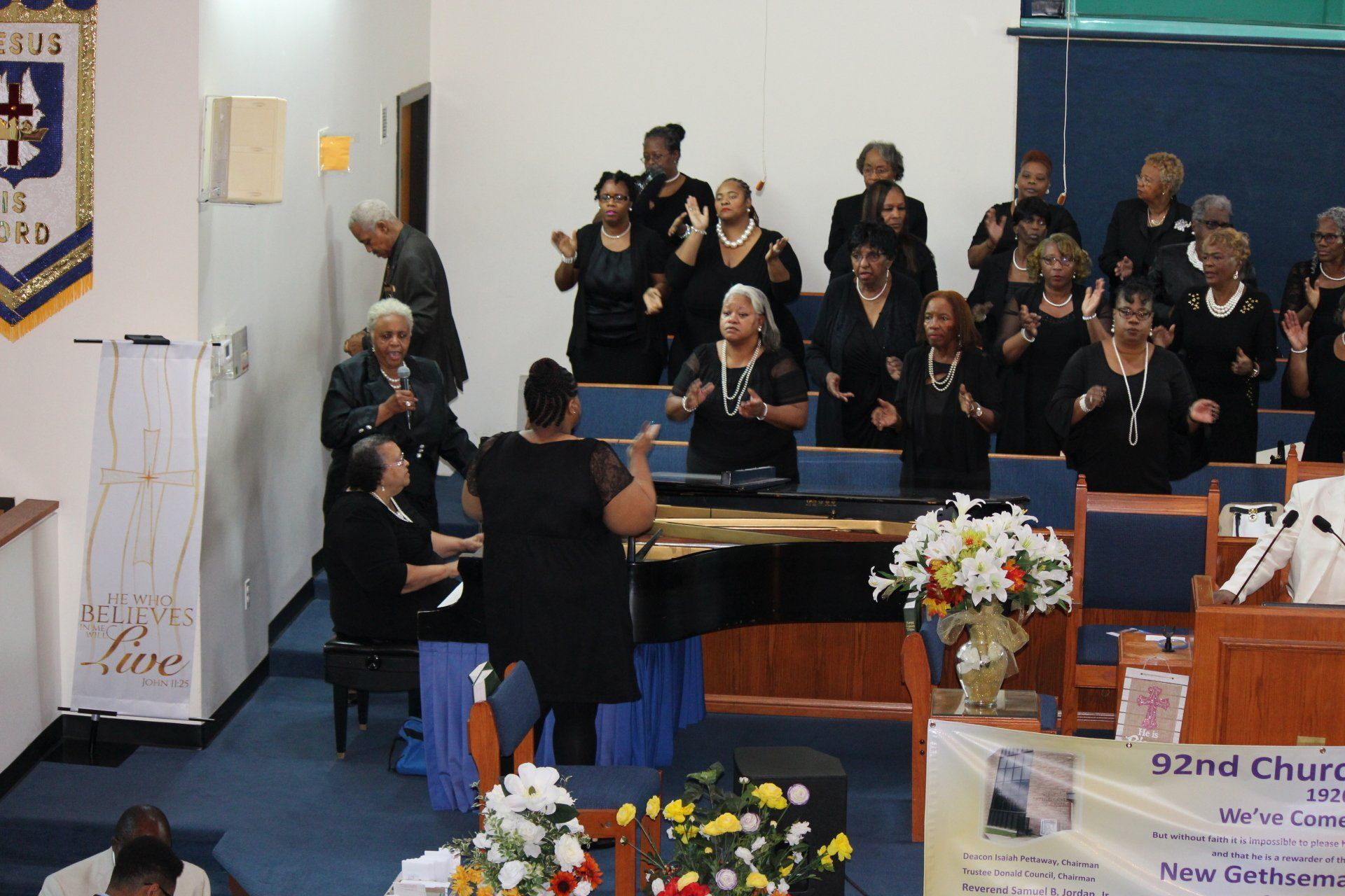 Church choir singing on stage, led by a conductor with raised arms, near a piano and floral arrangements.