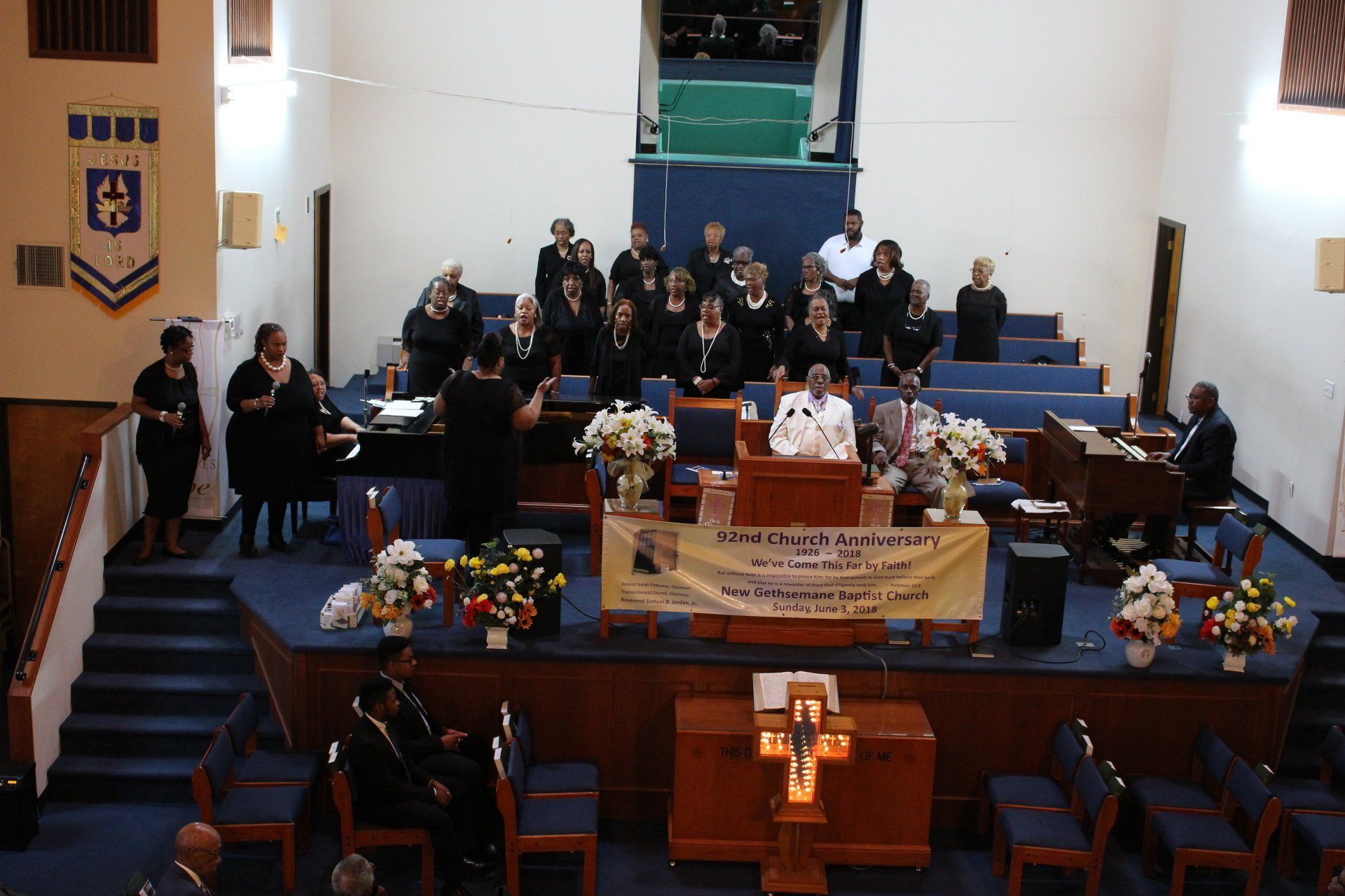 Church sanctuary with people seated on a stage behind a podium and floral arrangements, viewed from the back.