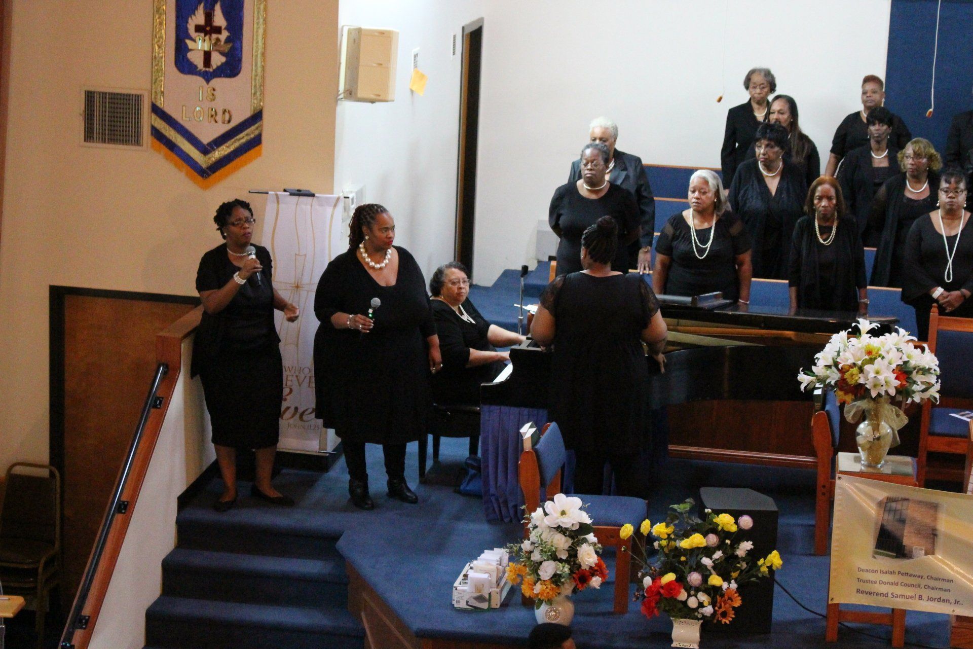 Choir singing in a church sanctuary, with a conductor, floral arrangements, and people seated behind them