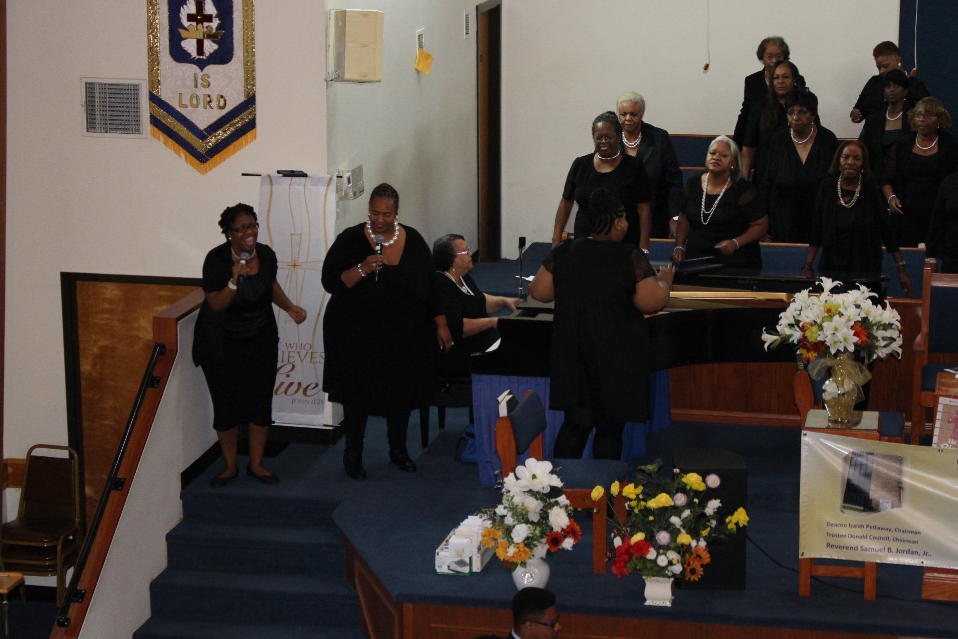 People standing on a church stage during a service, with flowers and a podium in front.