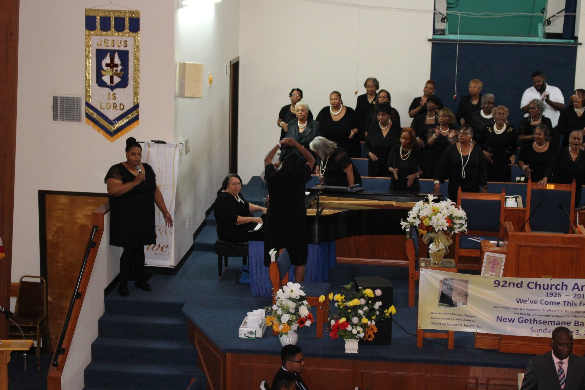 Choir singing on a church stage beside a speaker at a podium, with floral arrangements and banner.