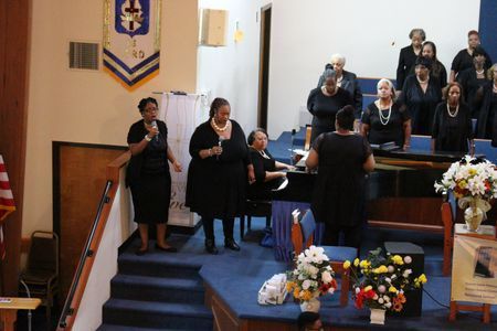 Choir singing in a church sanctuary beside a piano, with flowers and flags near the pulpit.