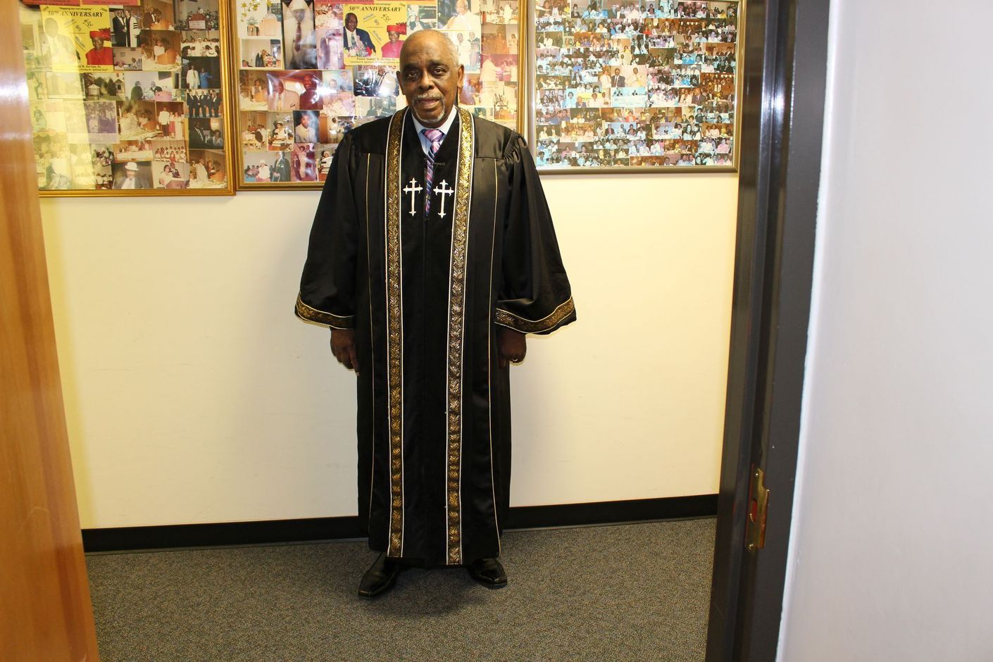 Pastor standing in a hallway wearing a black robe with gold trim, in front of colorful wall artwork.