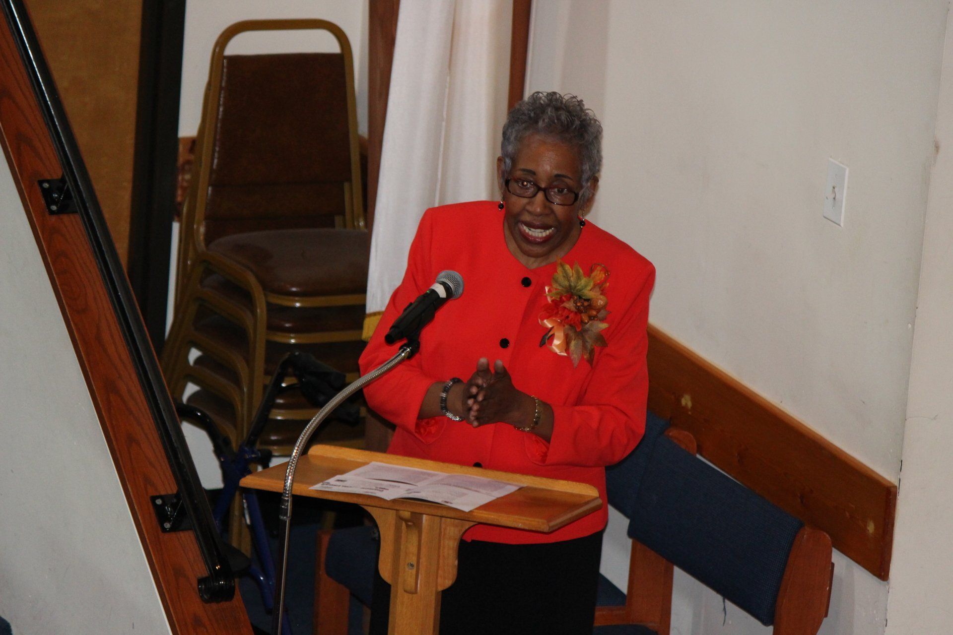 Woman in a red jacket speaking at a podium with a microphone in a small room