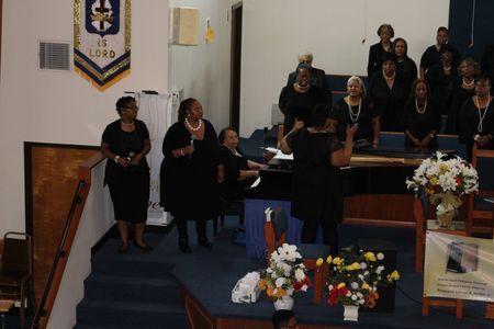 People gathered in a church auditorium, singing near a piano beside floral arrangements.