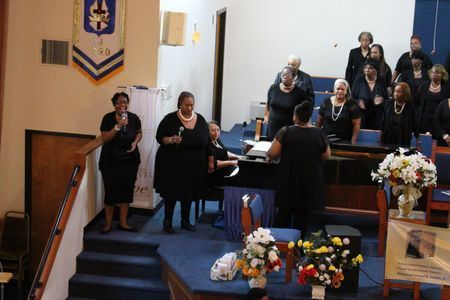 Choir singing in a church sanctuary beside floral arrangements and a wooden pulpit