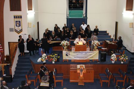 Church choir and speakers on stage during a ceremony, with floral arrangements and blue pews in front.