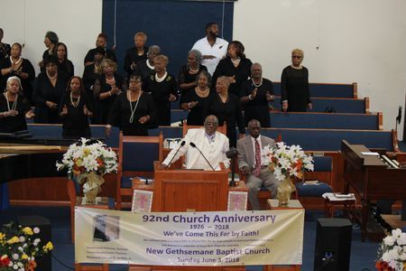Church choir and speakers at a 92nd anniversary service behind a floral banner on stage