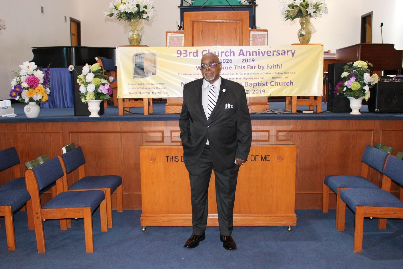 Man standing in a church sanctuary before a 30th anniversary banner and flowers, wearing a black suit.