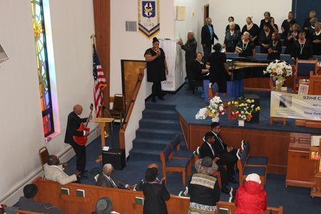 People gathered in a church sanctuary during a ceremony, with a speaker at the pulpit and choir on stage.