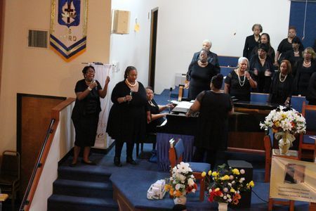 Choir singing in a church sanctuary, with a conductor facing them and floral arrangements near the pulpit.